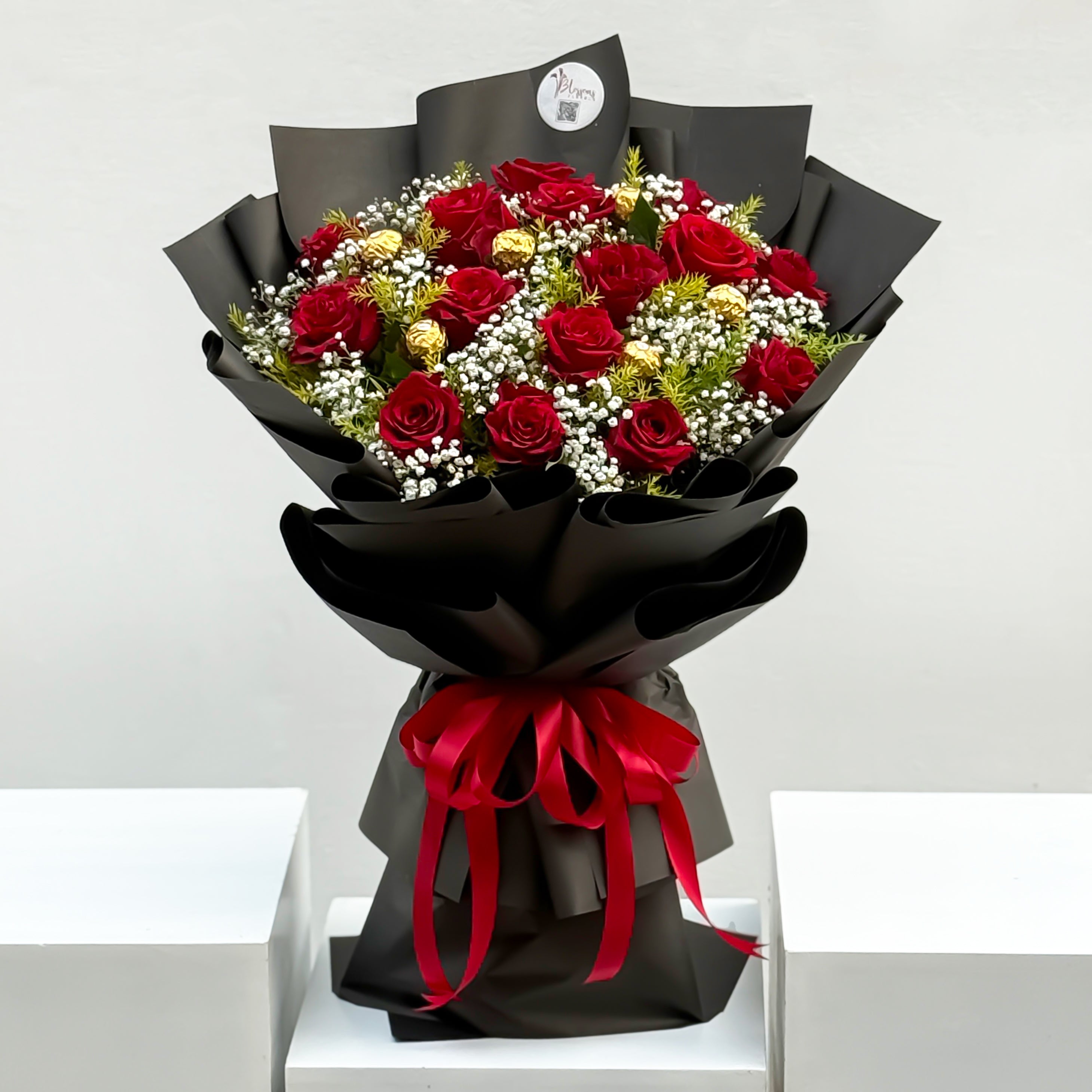Bouquet of red roses and greenery wrapped in black paper with a red ribbon on a white surface.