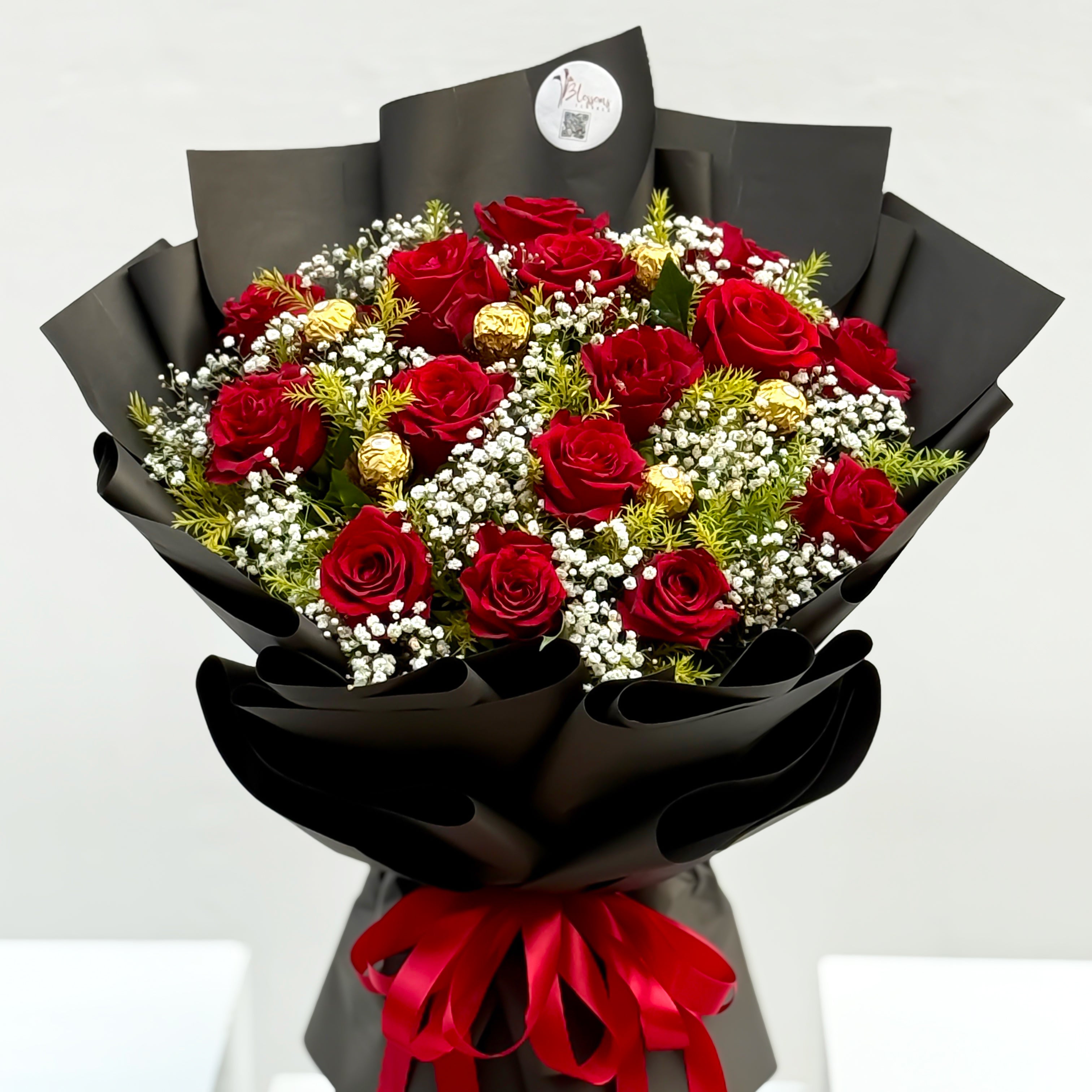 Bouquet of red roses with greenery and white flowers, wrapped in black paper with a red ribbon on a white background.
